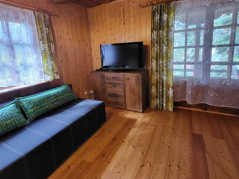 Rozkładana kanapa z zielonymi poduszkami i telewizor na drewnianej komodzie, duże okna z firankami